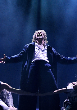 Zdjęcie ze spektaklu - główny bohater śpiewa na scenie w otoczeniu tancerzy.