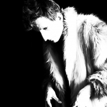 Czarno-białe zdjęcie z wysokim kontrastem. Osoba na zdjęciu ma założone futro, ma krótko obcięte kręcone włosy. Z rękawków wyłaniają się kościste dłonie z długimi, zaniedbanymi paznokciami