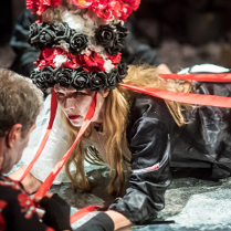 Zdjęcie ze spektaklu - kobieta z czepcem w biało-czerwono-białych kolorach na głowie, leżąca i patrzaca na mężczyznę, którego twarz jest niewidoczna.