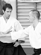 Dwóch mężczyzn w białych strojach do aikido trenuje sztuki walki w sali gimnastycznej.