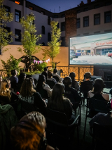 Grupa ludzi siedzi na krzesłach i ogląda film na ekranie plenerowym. Widzowie mają na uszach słuchawki, przez które słyszą dźwięk filmu. Na balkonie, na którym siedzą postawiono małe drzewka, a między nimi rozwieszono różowe girlandy.