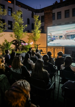 Grupa ludzi siedzi na krzesłach i ogląda film na ekranie plenerowym. Widzowie mają na uszach słuchawki, przez które słyszą dźwięk filmu. Na balkonie, na którym siedzą postawiono małe drzewka, a między nimi rozwieszono różowe girlandy.