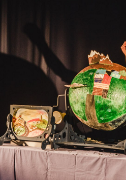 Dwie osoby w szaryych strojach na scenie teatralnej, stojące obok kartonowej scenografii.