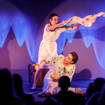 Dwie kobiety ubrane w ludowe sukienki występują na scenie. Jedna siedzi na ziemi, a druga nad nią macha kawałkiem materiału.