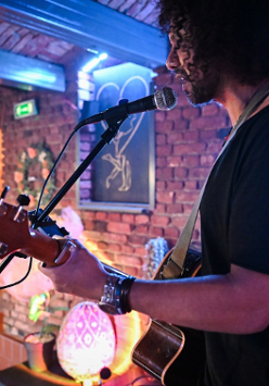 Mężczyzna z kręconymi włosami. Trzyma w rękach gitarę. Śpiewa do mikrofonu. W tle ceglana ściana z ozdobami.