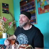 Tomasz Budzyński w czarnej czapce i ciemnej koszulce z gitarą w mieszkaniu. Po lewej zielona ściana, po prawej turkusowa. Na ścianie obrazy.