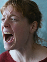 Zbliżenie na twarz krzyczącej kobiety. Kobieta ma rude włosy i czerwoną bluzkę. Stoi na tle tablicy w klasie.