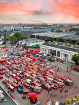 Widok z góry na Międzynarodowe Targi Poznańskie. Na placu głównym stoją zaparkowane wozy strażackie w kolorze czerwonym. Wokół nich gromadzą się ludzie, którzy oglądają pojazdy i robią zdjęcia. W oddali rozpościera się panorama Poznania.