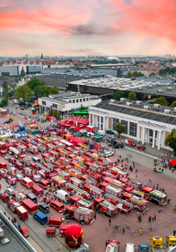 Widok z góry na Międzynarodowe Targi Poznańskie. Na placu głównym stoją zaparkowane wozy strażackie w kolorze czerwonym. Wokół nich gromadzą się ludzie, którzy oglądają pojazdy i robią zdjęcia. W oddali rozpościera się panorama Poznania.