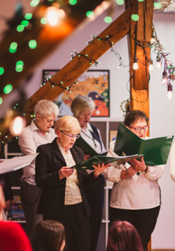 Grupa śpiewającycj osób w udekorowanym świątecznie pomieszczeniu.