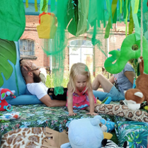 Mała dziewczynka bawi się na kolorowej macie, na której leżą różne pluszaki. Za dziewczyną siedzą rodzice, którzy rozmawiają ze sobą.