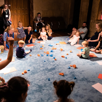 Zdjęcie sali, w której na podlodze pokrytej jasno niebieskim materiałem siedzą dzieci, przy nich rodzice. Na środku rozrzucone niebieskie i pomarańczowe zgniecione kawałki papieru.