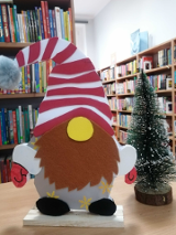 Na pierwszym planie świąteczny skrzat z brodą i czerwoną czapeczką. Obok ozdobna choinka. W tle regały z książkami.