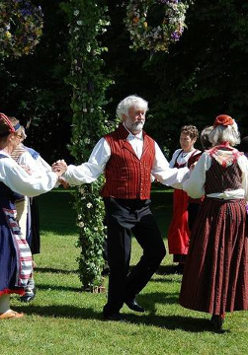 Na zdjęciu widać grupę ludzi, głównie kobiet, ubranych w tradycyjne skandynawskie stroje ludowe. Tańczą w kole wokół maja w parku. Kobiety noszą jaskrawo kolorowe suknie z misterną haftowaną ozdobą, a mężczyźni mają białe koszule z kamizelkami. Maj jest udekorowany kwiatami, wstążkami i zielenią. Scena wydaje się być uroczysta i radosna.