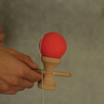 Zdjęcie, ręka trzyma drewniane urządzenie, do którego przymocowano za pomocą sznurka czerwoną piłkę.