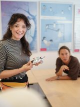 Zdjęcie przedstawia uśmiechniętą kobietę, stojącą pod tablicą w pomieszczeniu przypominającym małą salę lekcyjną.