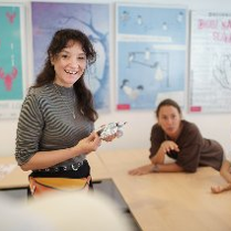 Zdjęcie przedstawia uśmiechniętą kobietę, stojącą pod tablicą w pomieszczeniu przypominającym małą salę lekcyjną.