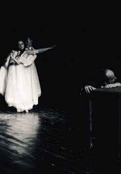 Na scenie widoczny mężczyzna opierający głowę i ręce o stolik, w tle stoją dwie kobiety w długich białych sukniach, unoszą ręce do góry