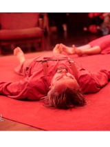 Na podłodze, na czerwonym dywanie leży kilka osób, zwróconych bosymi strojami do środka. Mają również czerwone stroje, a całe zdjęcie ma czerwoną poświatę.