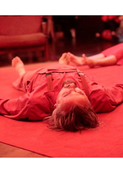 Na podłodze, na czerwonym dywanie leży kilka osób, zwróconych bosymi strojami do środka. Mają również czerwone stroje, a całe zdjęcie ma czerwoną poświatę.