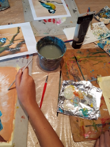 Farby, pędzle rozrzucone na stole. Miedzy nimi malunki kolorowych ptaktów.