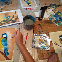 Farby, pędzle rozrzucone na stole. Miedzy nimi malunki kolorowych ptaktów.