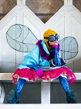 Kobieta przebrana w kolorowe ciuchy za muchę siedząca na białej ławie.