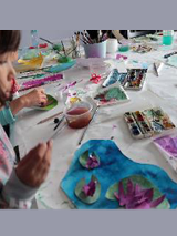 Stół z farbami i przyborami malarskimi, po lewej stronie dziewczynka z pędzelkiem