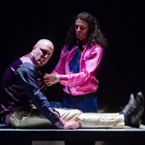 Na zdjęciu dwóch aktorów na czarnym tle. Jeden siedzi z wyprostowanymi nogami, drugi klęczy obok.