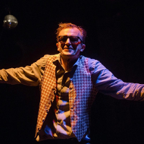 Na zdjęciu na czarnym tle, aktor w ciemnych okularach z rozłożonymi ramionami.