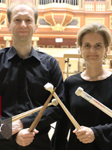 Na zdjęciu dwoje muzyków z pałeczkami perkusyjnymi. Stoją na scenie auli UAM, w tle organy.