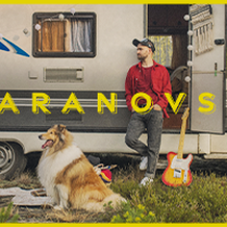 Na zdjęciu mężczyzna opierający się o przyczepę campingową. W otwartych drzwiach przyczepy stoi gitara. Przed przyczepą leży pies.