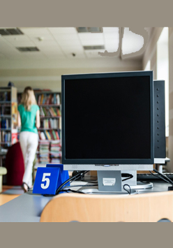 Na zdjęciu pomieszczenie biblioteczne. Na pierwszym planie stolik z komputerem, w tle regały z książkami.
