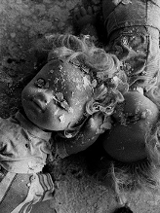 Czarno-białe zdjęcie przysypanych fragmentami pokruszonego tynku dwóch lalek, leżących na zniszczonym blacie stołu.