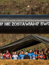 Grupa osób w strojach sportowych. Napis "Nigdy nie zostawiamy swoich". Obok stogi siana i inne przeszkody.
