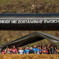 Grupa osób w strojach sportowych. Napis "Nigdy nie zostawiamy swoich". Obok stogi siana i inne przeszkody.