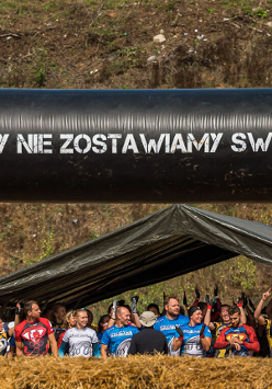 Grupa osób w strojach sportowych. Napis "Nigdy nie zostawiamy swoich". Obok stogi siana i inne przeszkody.