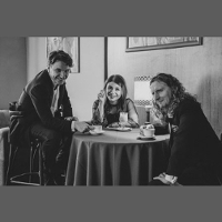 Czarno-biała fotografia. Na zdjęciu dwóch mężczyzn i kobieta. Siedzą przy stoliku kawiarnianym, w eleganckich strojach. Patrzą w obiektyw aparatu i się uśmiechają. Na stole filiżanki z kawą.