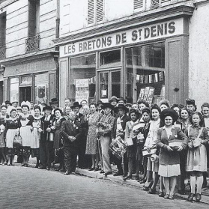 Czarno-białe zdjęcie, ludzie stojący przed budynkiem z napisem "Les Bretons de St Denis"