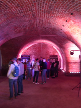 Grupa ludzi w podziemiach kaponiery, czytają plakaty rozstawione w pomieszczeniu.