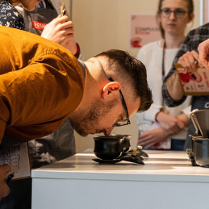 Zdjęcie przedstawiające mężczyznę schylonego i wąchającego napój w filiżance, obserwuje go grupa ludzi