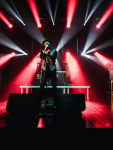 Na zdjęciu artysta śpiewający na scenie na tle czerwono białych świateł.