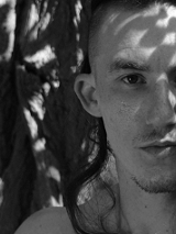 Czarno białe zdjęcie przedstawia twarz mężczyzny z lekkim zarostem i długimi włosami na tle drzewa.