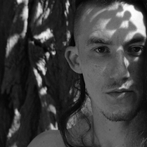 Czarno białe zdjęcie przedstawia twarz mężczyzny z lekkim zarostem i długimi włosami na tle drzewa.