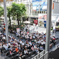 zdjęcie z targów, na którym widoczni są ludzie siedzący przed sceną przed budynkiem MTP