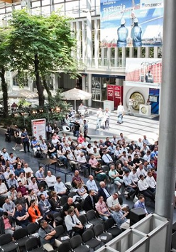 zdjęcie z targów, na którym widoczni są ludzie siedzący przed sceną przed budynkiem MTP