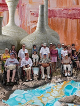 Zdjęcie przedstawia grupę osób z niepełnosprawnościami znajdujących się na pustkowiu. W tle widoczne są olbrzymie wazy, widoczne są także ślady farby.