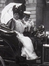 kadr z czarno-białego filmu "Kobiety za kierownicą"; na siedzeniu samochodu prowadzonego przez szofera siedzi kobieta w białej sukni trzymająca w ręku parasol