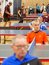 zdjęcie z turnieju tenisa stołowego; na zdjęciu widoczni grający zawodnicy i przypatrujący się grze sędziowie