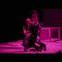 Aktorka grająca rolę George Sand trzyma przed sobą lalkę wyobrażającą Chopina. Ciemne tło, na podłodze różowa tkanina.